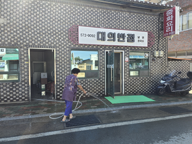#1. 250804 보도사진2(1일 대의반점 배영자 사장이 가게 앞 물을 뿌리며 장사 개시를 준비하고 있다)